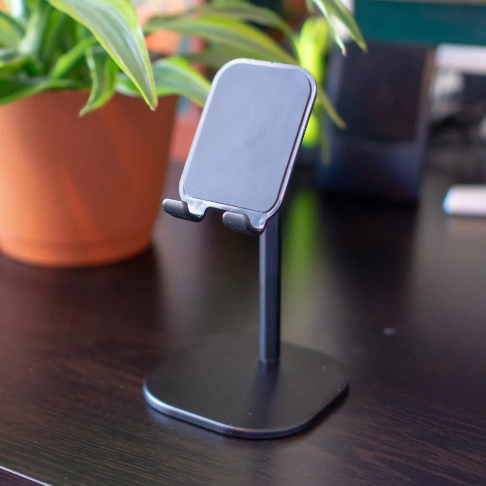 A phone in a rigid aluminum desk stand on a wooden desk, next to a potted plant.